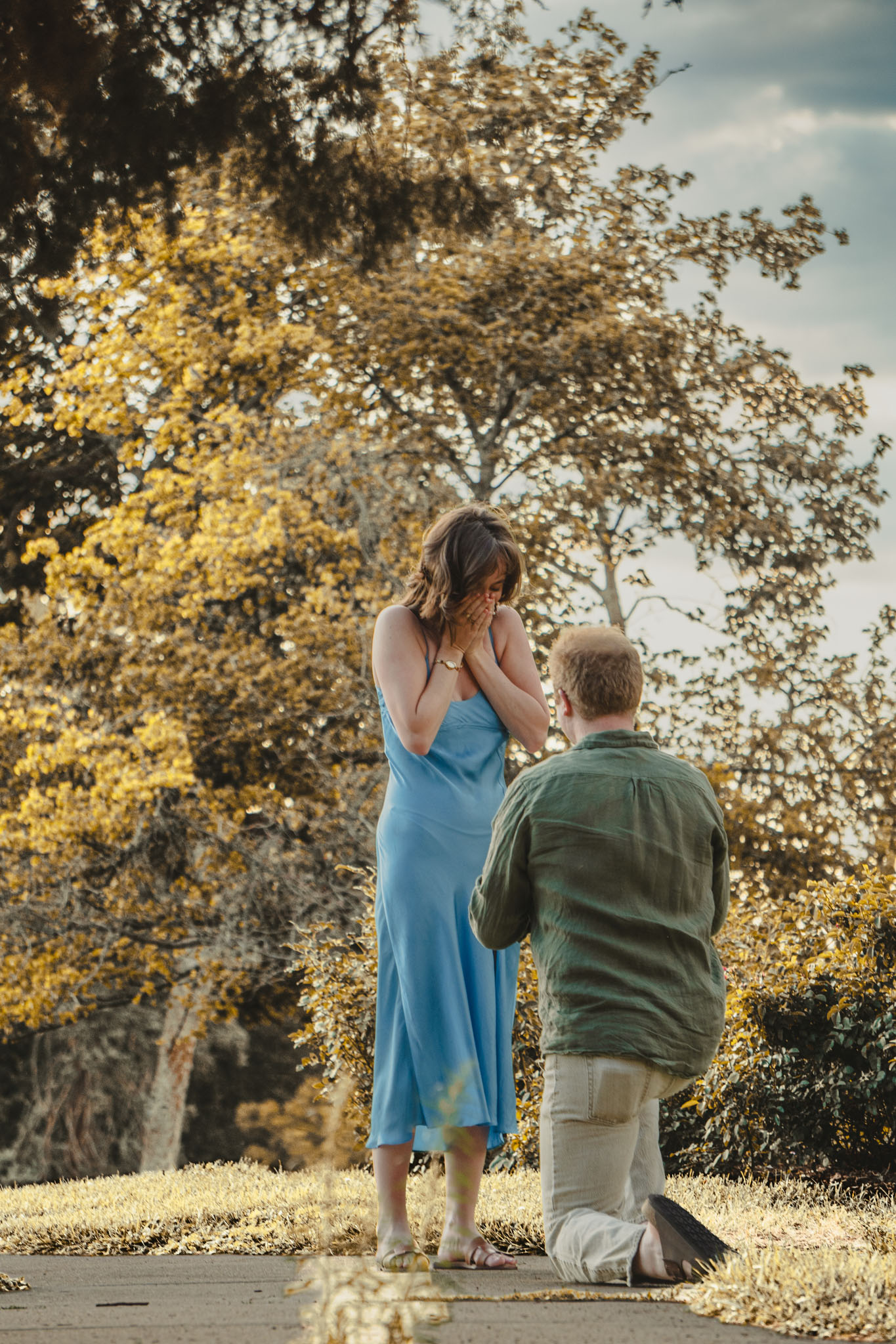 One partner kneeling during an outdoor proposal as the other covers their mouth.
