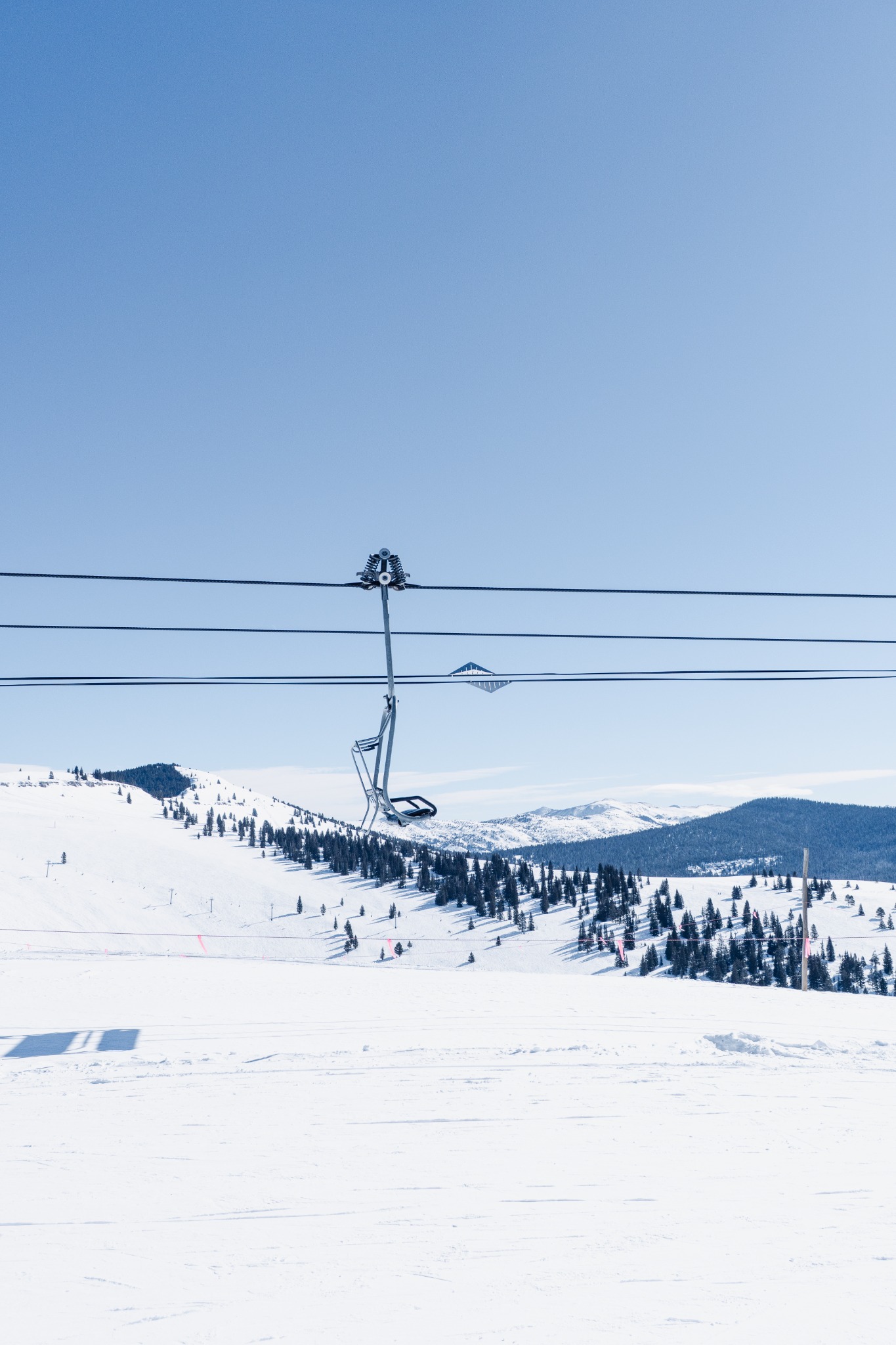 An empty ski chair suspended above snowy slopes and evergreen trees under a pale blue sky.
