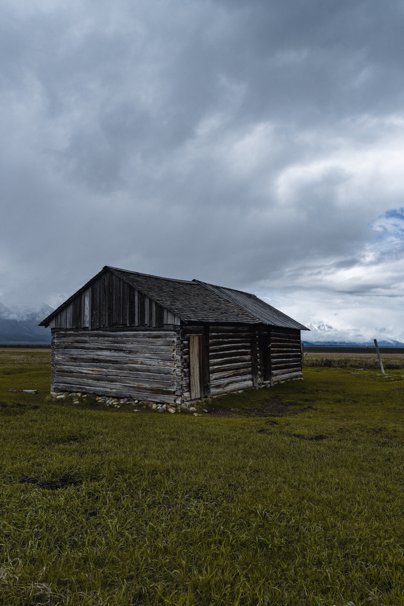 A weathered wooden cabin in a green meadow under low gray clouds with mountains in the distance.