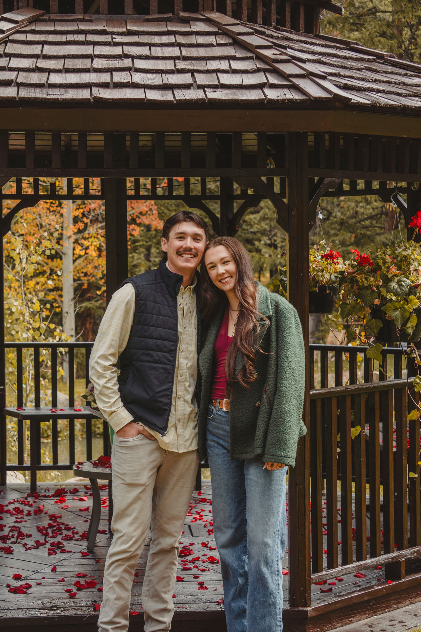 Couple standing together and smiling beneath a wooden gazebo.