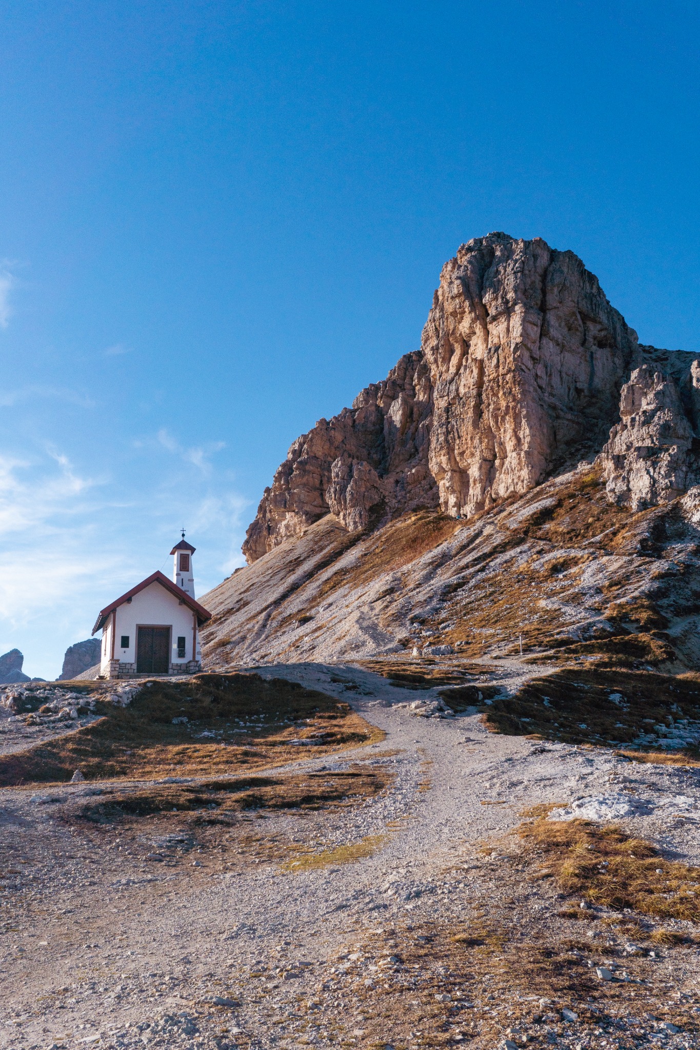 A small white chapel beside a rocky mountain trail below a tall sunlit cliff and blue sky.