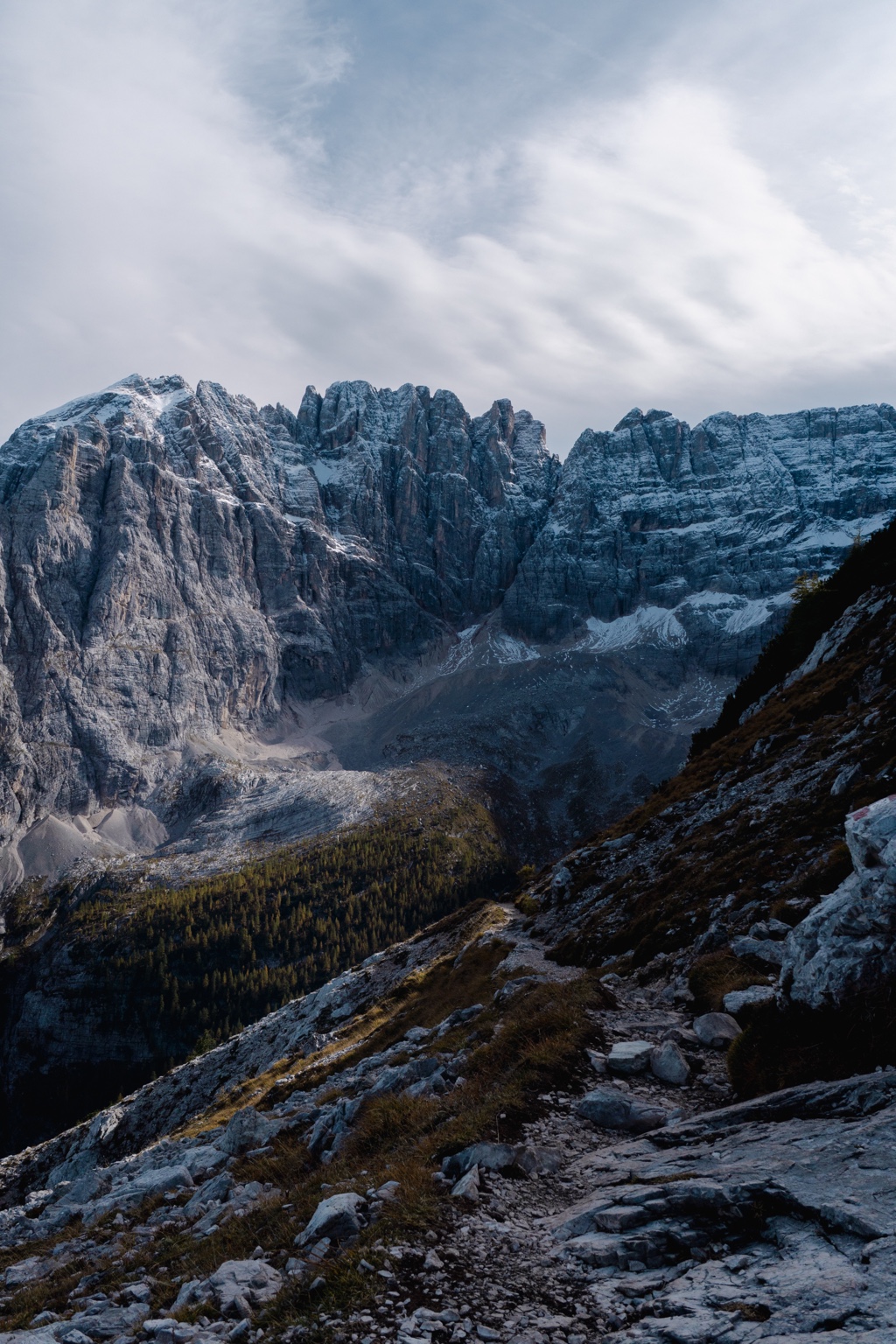 Jagged snow-dusted rock towers above a winding mountain path and a forested valley.