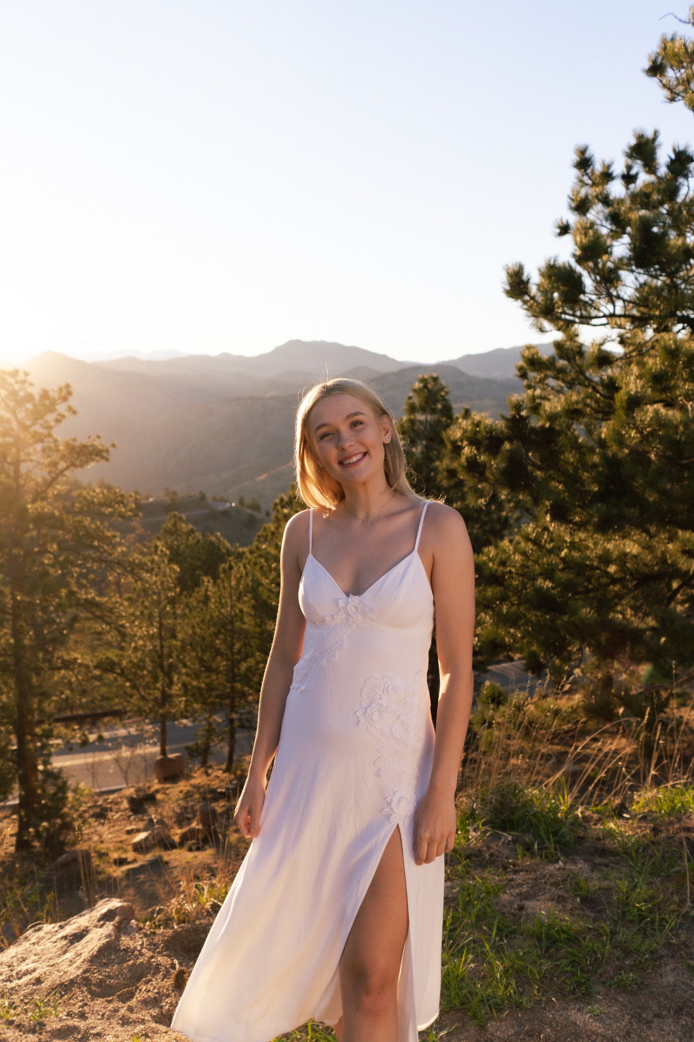 Outdoor portrait in a white dress with pine trees and mountains in warm evening light.