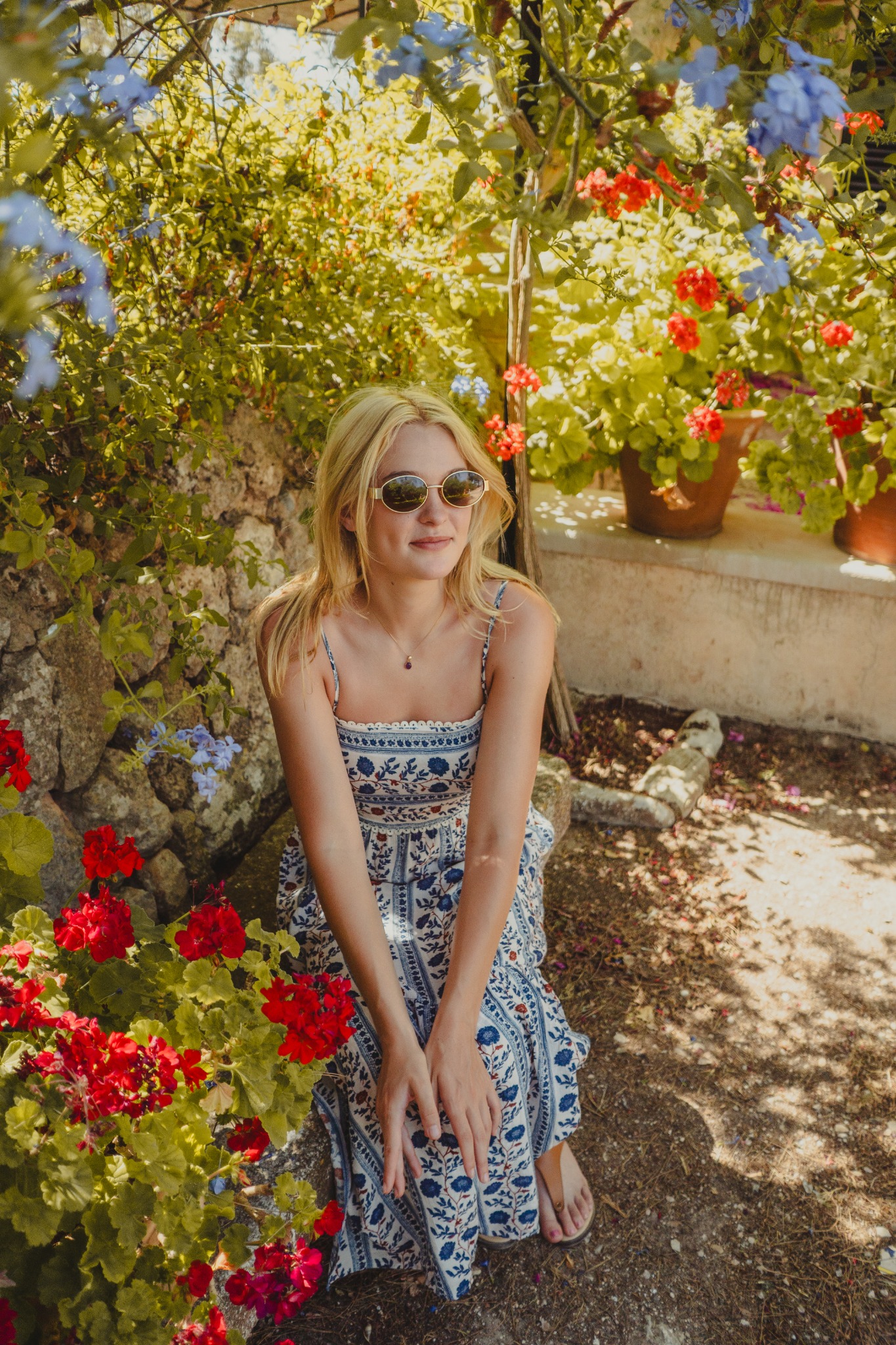 Woman in a blue patterned dress and sunglasses seated among red and blue garden flowers.