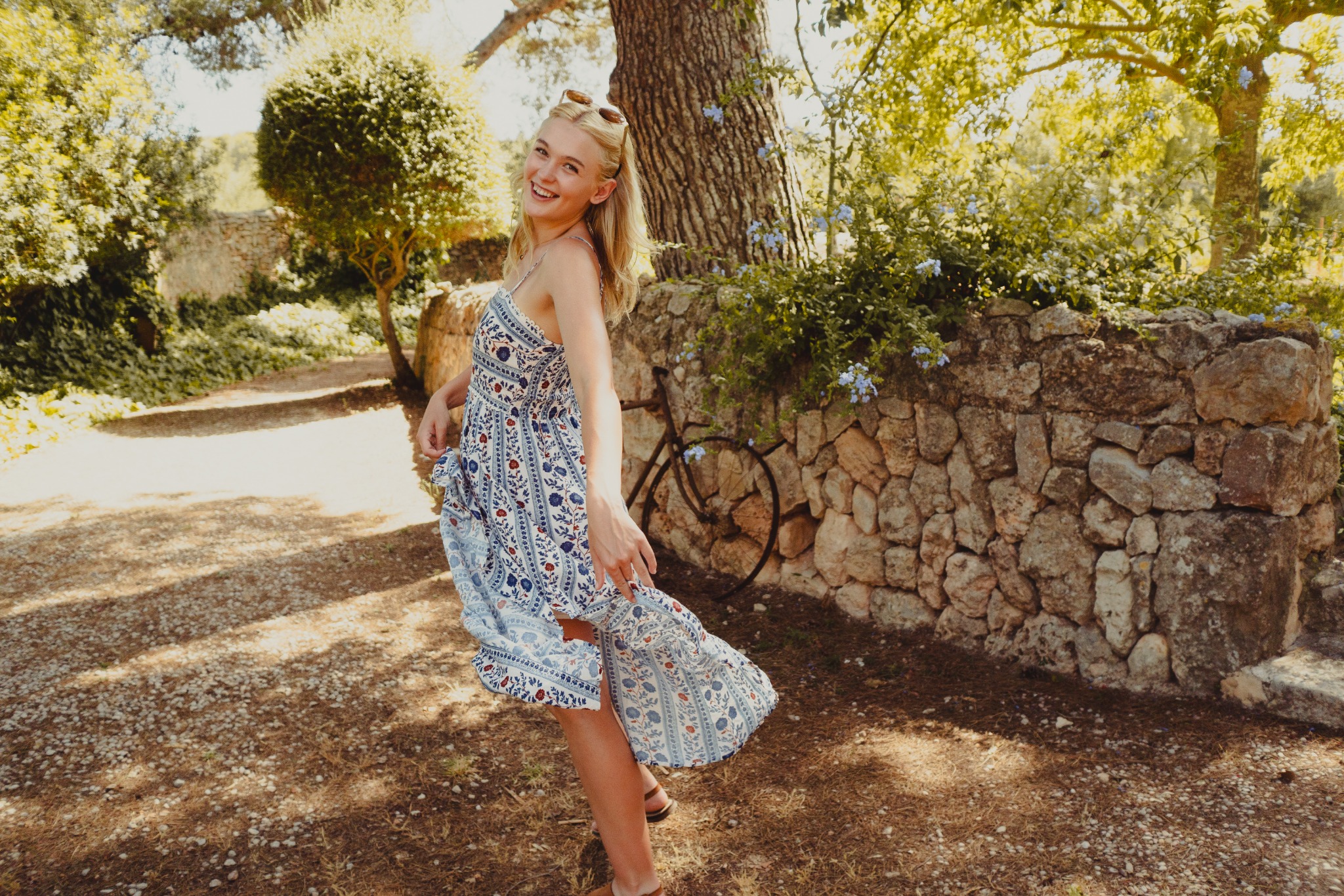 Smiling woman in a blue patterned dress beside a stone garden wall and bicycle.