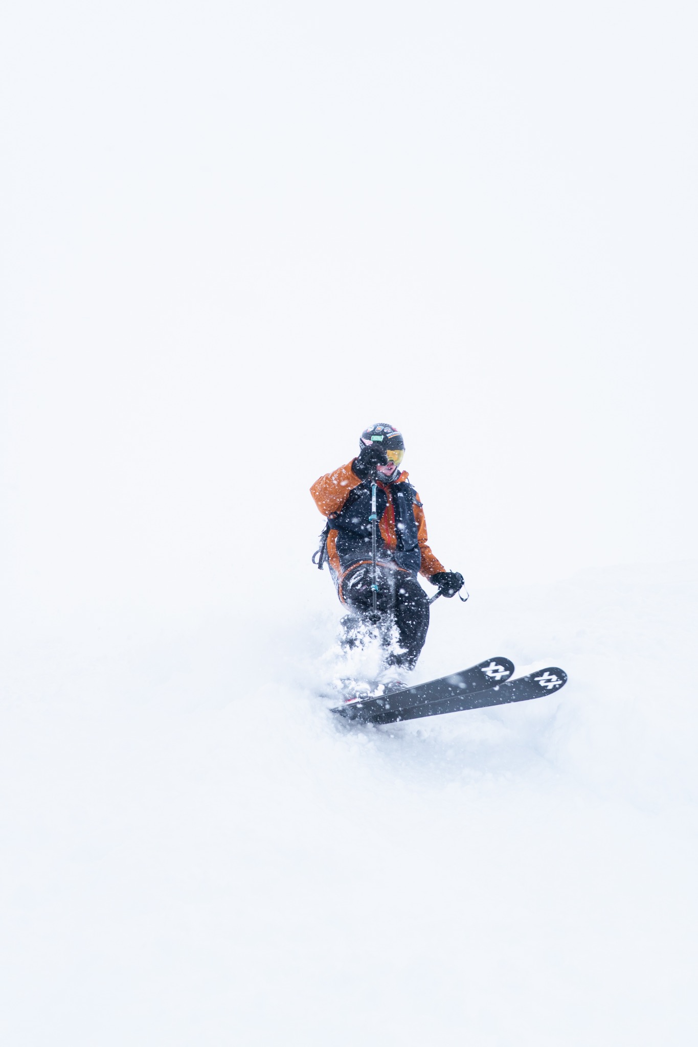 Skier in an orange and black jacket moving through fresh powder in falling snow.